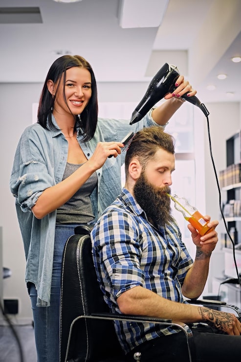 Kostenlose Foto brünette friseurin, die eine frisur mit haartrockner des bärtigen männerkunden in einem salon macht.