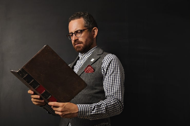 Kostenlose Foto ernsthafter männlicher lehrer in tweedweste und kariertem hemd liest ein großes altes buch an einer tafel