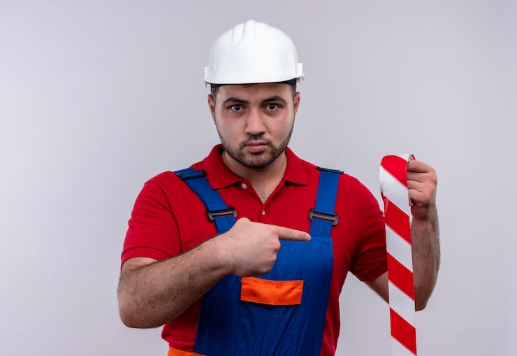 Kostenlose Foto junger baumeistermann in der bauuniform und im sicherheitshelm, der klebeband hält, das mit zeigefinger auf ihn mit ernstem gesicht zeigt