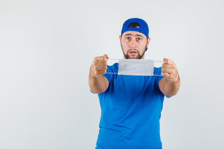 Kostenlose Foto junger mann, der medizinische maske im blauen t-shirt und in der kappe hält und vorsichtig schaut