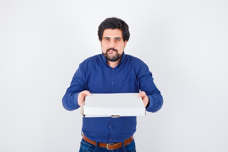 Kostenlose Foto junger mann im hemd, jeans, die geschlossene pizzaschachtel hält und hungrig aussieht, vorderansicht.