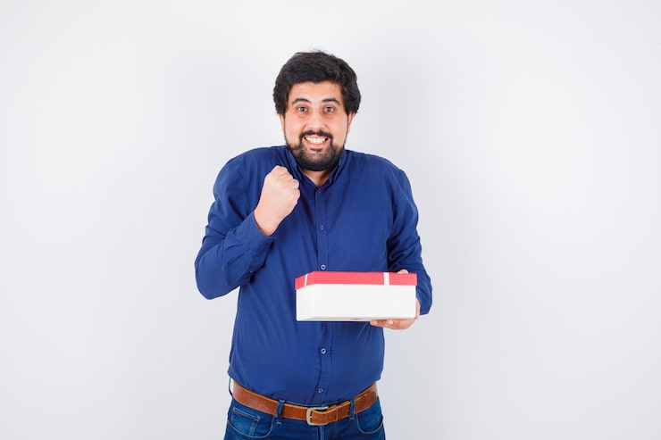 Kostenlose Foto junger mann in blauem hemd und jeans mit geschenkbox und geballter faust und glücklich aussehend, vorderansicht.