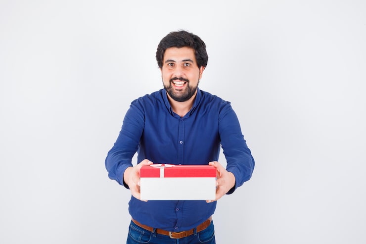 Kostenlose Foto junger mann präsentiert geschenkbox mit beiden händen in blauem hemd und jeans und sieht optimistisch aus. vorderansicht.