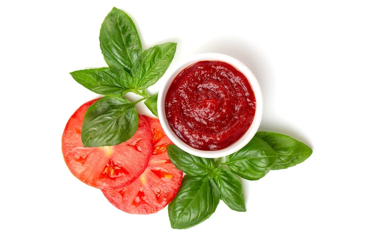 Foto ketchup (pasta de tomate) en una cacerola con hojas de albahaca y rodajas de tomate sobre un fondo blanco.