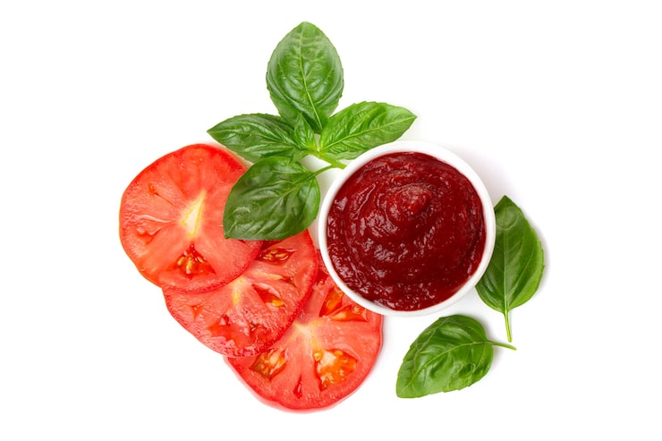 Foto ketchup (pasta de tomate) en una cacerola con hojas de albahaca y rodajas de tomate sobre un fondo blanco.