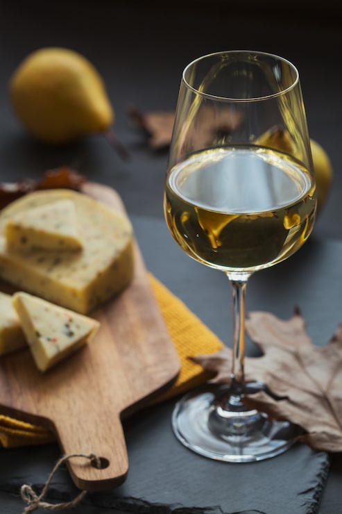 Foto um copo de vinho branco servido com queijo em uma tábua em fundo escuro