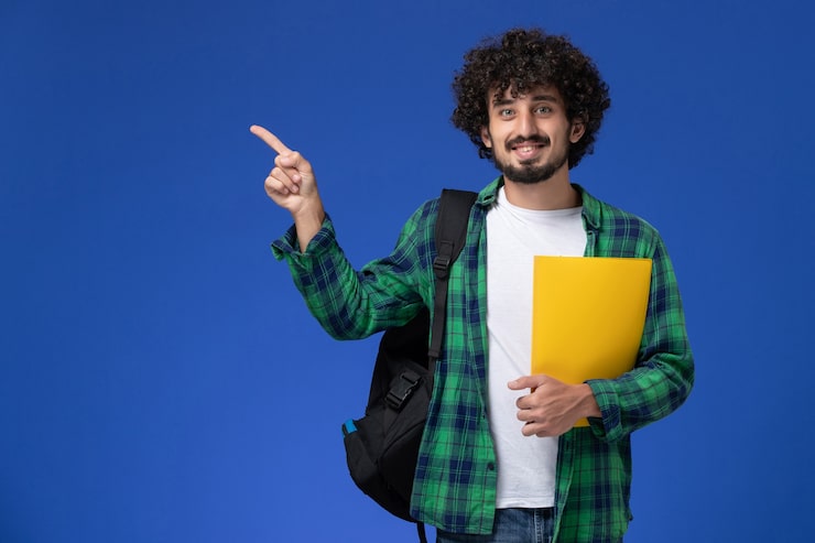 Gratis Bild framifrån vy av manlig student i grönrutig skjorta med svart ryggsäck som håller filer och ler mot den blå väggen