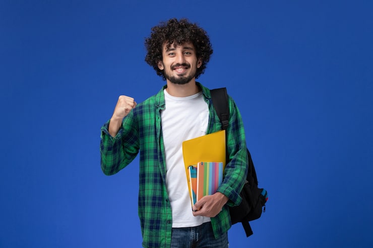Gratis Bild framifrånvy av manlig student som bär svart ryggsäck och håller kopieringsböcker och filer mot en blå vägg