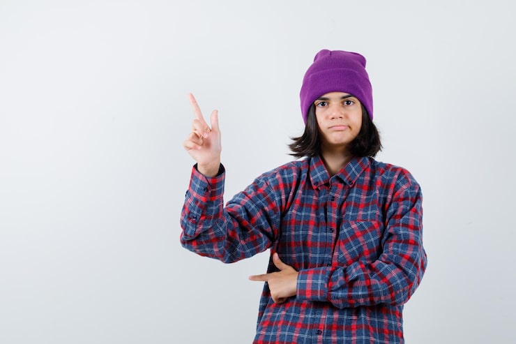 Gratis Foto teenage pige i ternet skjorte og hue peger opad og ser forsigtig ud