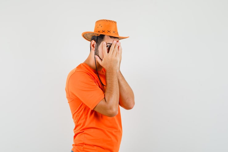 Gratis Bild ung man i orange t-shirt och hatt som tittar genom fingrarna och ser rädd ut, framifrån.