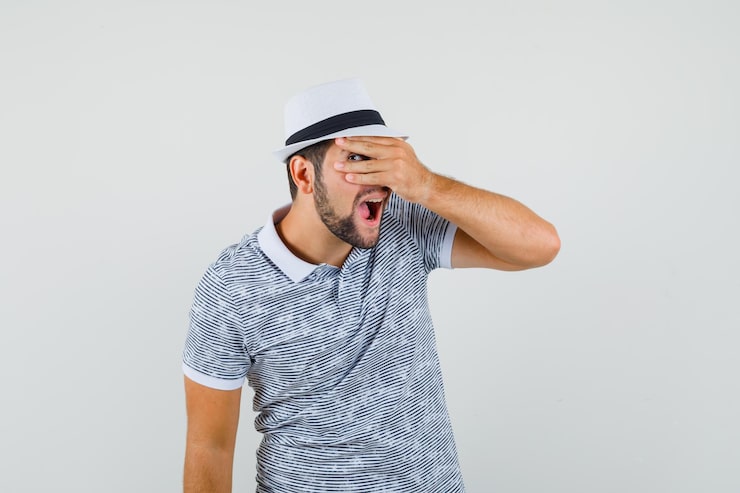 Gratis Bild ung man i t-shirt och hatt som tittar genom fingrarna och ser nyfiken ut, framifrån.