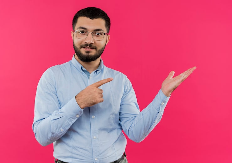 Gratis Foto ung, skægget mand i briller og blå skjorte, der præsenterer noget med armen og peger med pegefingeren til siden og smiler selvsikkert