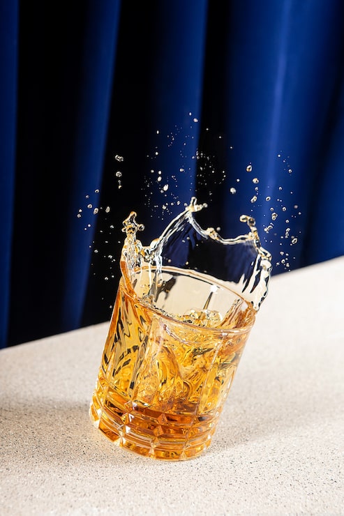 Gratis Bild vertikal bild av stänkande whisky i ett glas med en blå gardin i bakgrunden