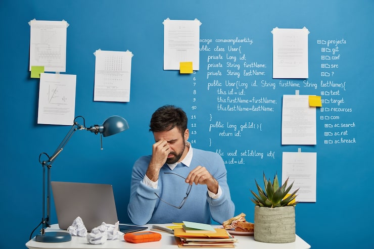 Photo gratuit un homme d'affaires épuisé se frotte le nez, enlève ses lunettes, souffre de fatigue oculaire et de maux de tête, a des problèmes au travail, est assis dans un espace de coworking avec un ordinateur portable, un mur bleu avec des notes écrites.