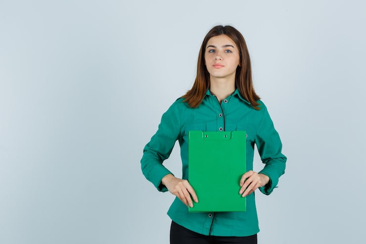 Photo gratuit jeune fille tenant le presse-papiers à deux mains en chemisier vert, pantalon noir et à la recherche de sérieux. vue de face.