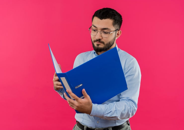 Photo gratuit jeune homme barbu à lunettes et chemise bleue regardant dossier avec visage sérieux