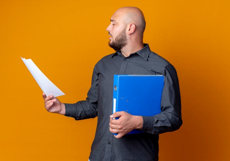Photo gratuit jeune homme de centre d'appels chauve tournant la tête à l'autre avec les yeux fermés tenant le dossier et le document isolé sur fond orange