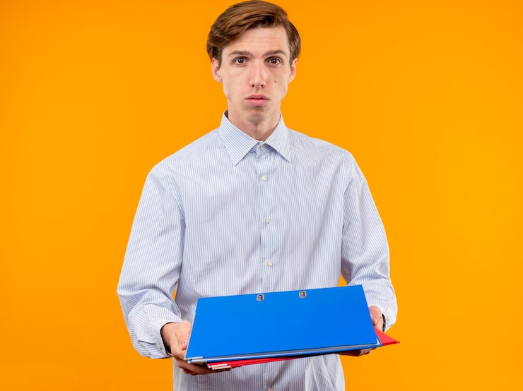 Photo gratuit jeune homme en chemise blanche tenant des dossiers regardant la caméra avec un visage sérieux debout sur fond orange