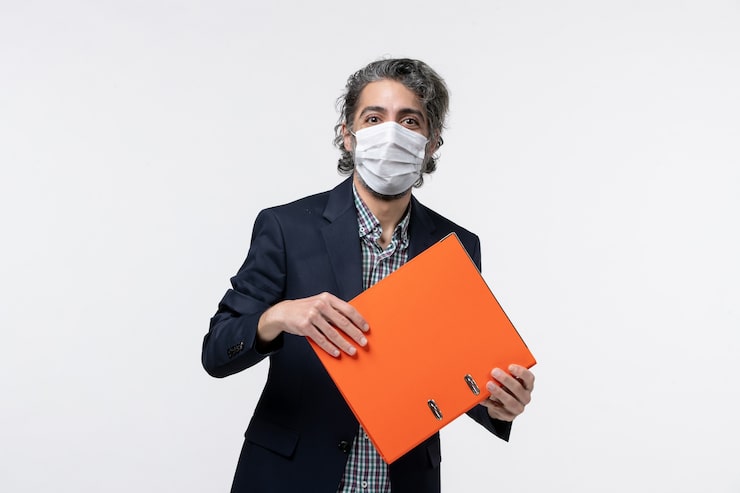 Photo gratuit souriant jeune homme en costume et tenant ses documents portant son masque chirurgical sur une surface blanche