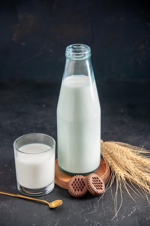 Photo gratuit vue verticale d'un lait frais dans une bouteille et des biscuits en verre et une cuillère dorée sur de petites pointes de plateau en bois marron sur fond de vague noire