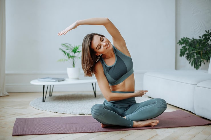 Bild leende ung brunett kvinna i en träningsoverall på en fitnessmatta hemma i vardagsrummet gör stretc