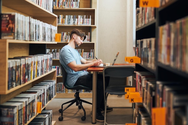 Studerende, der lærer i biblioteket, ung mand, der forbereder sig til test på bærbar computer, mand, der lytter til online kursus
