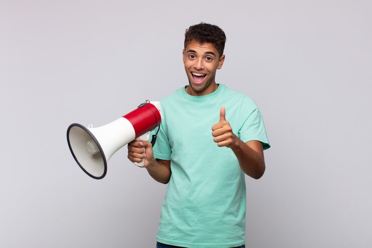 Foto ung mand med en megafon, der føler sig stolt, ubekymret, selvsikker og glad, smiler positivt med tommelfingrene opad