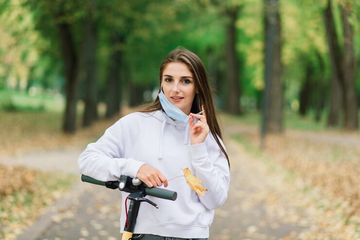 Foto toevallige kaukasische vrouw die een beschermend gezichtsmasker draagt, rijdt op een stedelijke elektrische scooter in het stadspark tijdens een covid-pandemie. stedelijk mobiliteitsconcept.