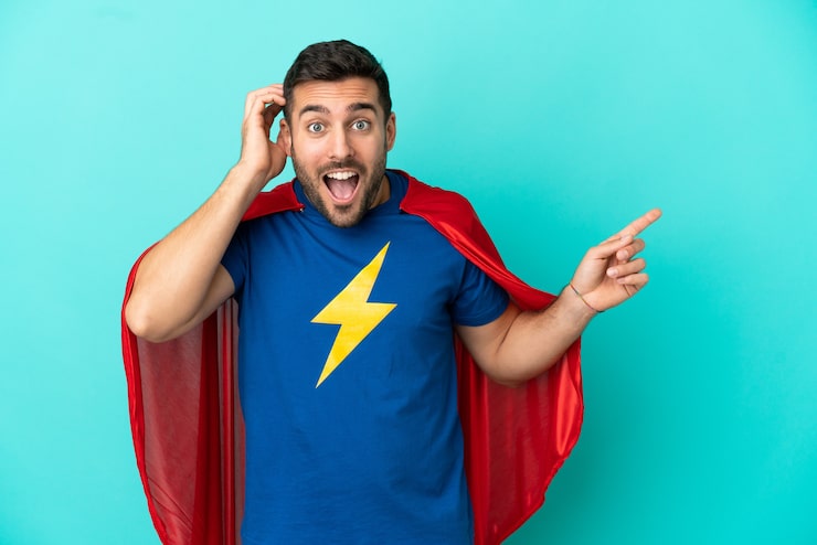 Zdjęcie super hero kaukaski mężczyzna na białym tle na niebieskim tle zaskoczony i wskazujący palcem na bok