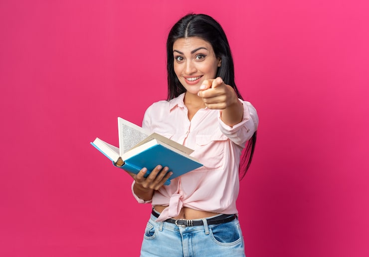 Ücretsiz Fotoğraf gündelik kıyafetler içinde kitap tutan, neşeyle gülümseyen, mutlu ve pozitif genç güzel kadın, pembe duvar önünde işaret parmağıyla işaret ediyor.
