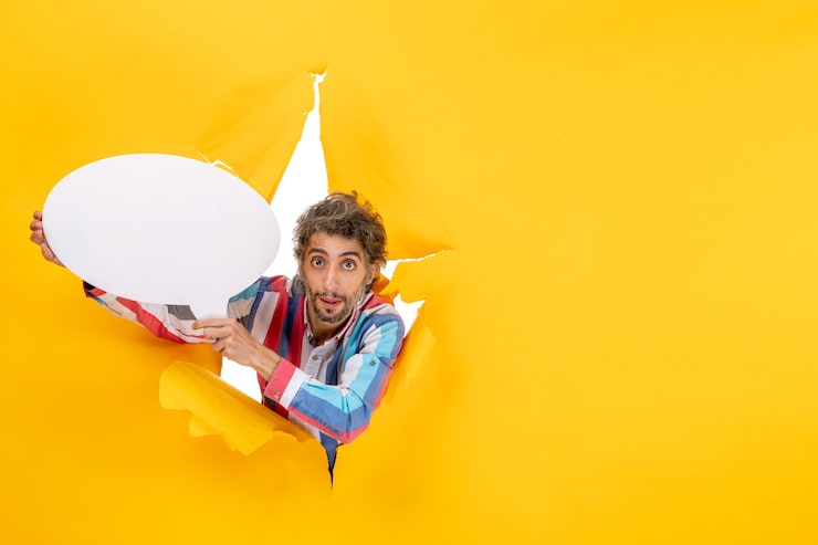 Ücretsiz Fotoğraf şaşırmış genç adam, sarı kağıttaki yırtık bir delikte kamera için beyaz balon tutarak poz veriyor