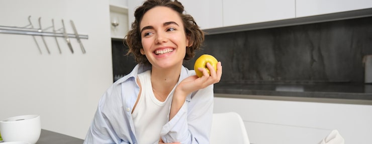 Gratis Foto afbeelding van brunette vrouw lachen glimlachend terwijl het eten van een appel in de keuken zit thuis met een