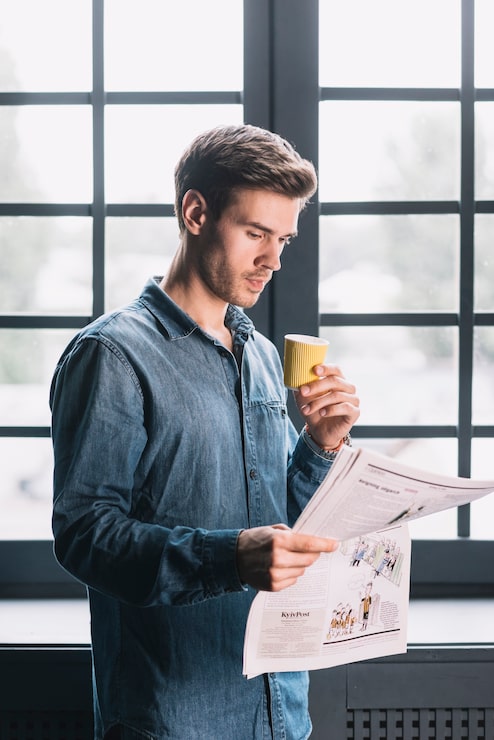 Gratis Foto jonge man permanent in de buurt van het venster met wegwerp koffiekopje krant lezen