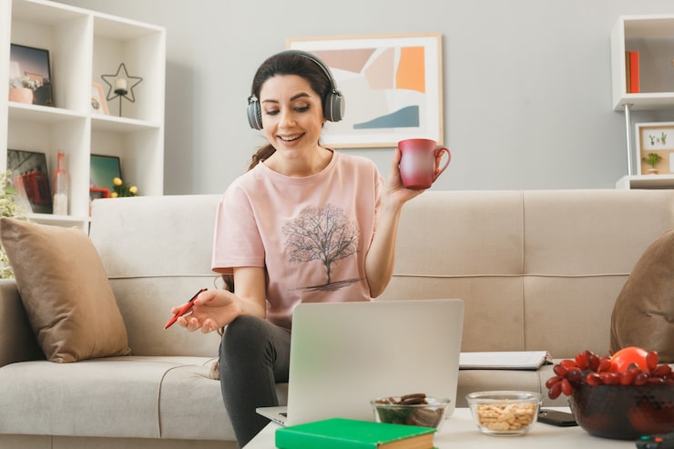 Gratis Foto jonge vrouw die een kopje thee vasthoudt met een pen die een koptelefoon draagt, gebruikte een laptop die op de bank zat achter de salontafel in de woonkamer