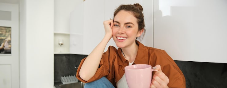 Gratis Foto mooi jong vrouwelijk model met een roze beker zit in de keuken en glimlacht naar de camera vrouw