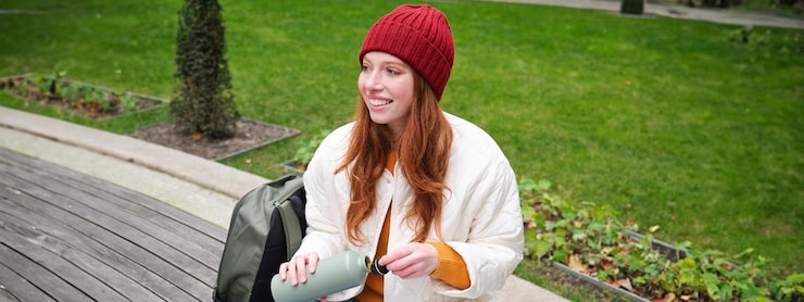 Gratis Foto portret van een jonge roodharige vrouw met een hoed die op een bank zit en een thermosfles vasthoudt en warme dranken drinkt