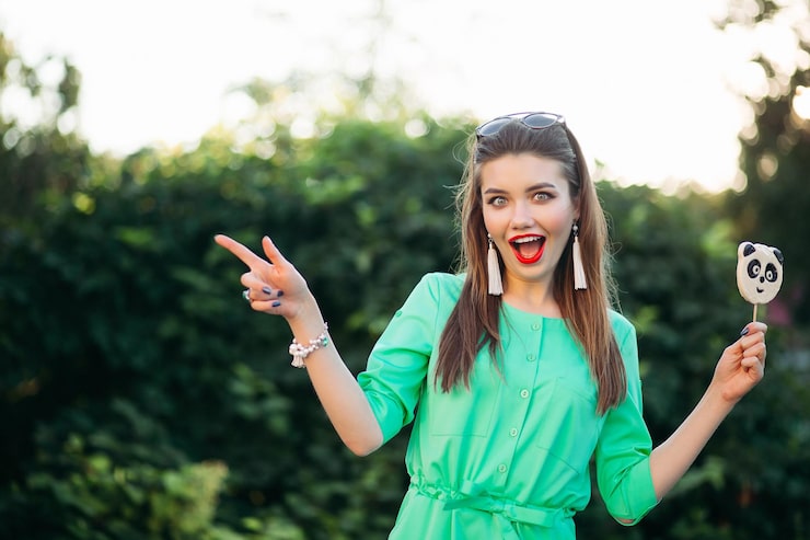 Gratis Foto portret van emotioneel en mooi brunette meisje in groene jurk, met snoep als panda op stok en verrast schreeuwen. mooie en stijlvolle vrouw met rode lippen, lang haar dat naar de zijkant wijst.
